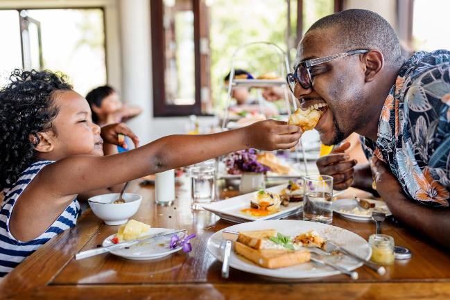 Stock image of a child sharing a bite of her food with her father.