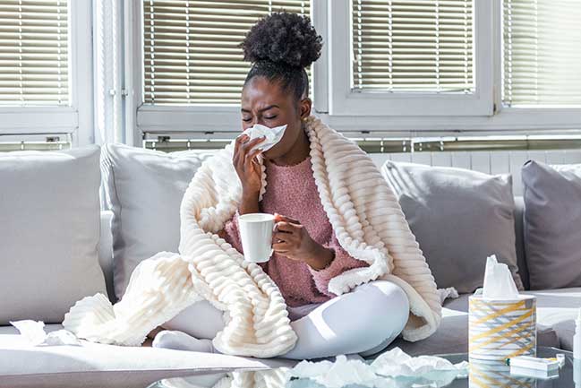 A woman sick with the flu, sitting on the couch and blowing her nose.