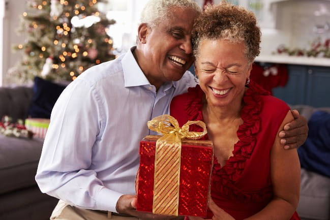 A couple smiles while holding a wrapped gift; a lit Christmas tree is scene in the background.