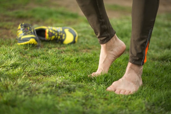 Barefoot person standing on grass next to athletic shoes.