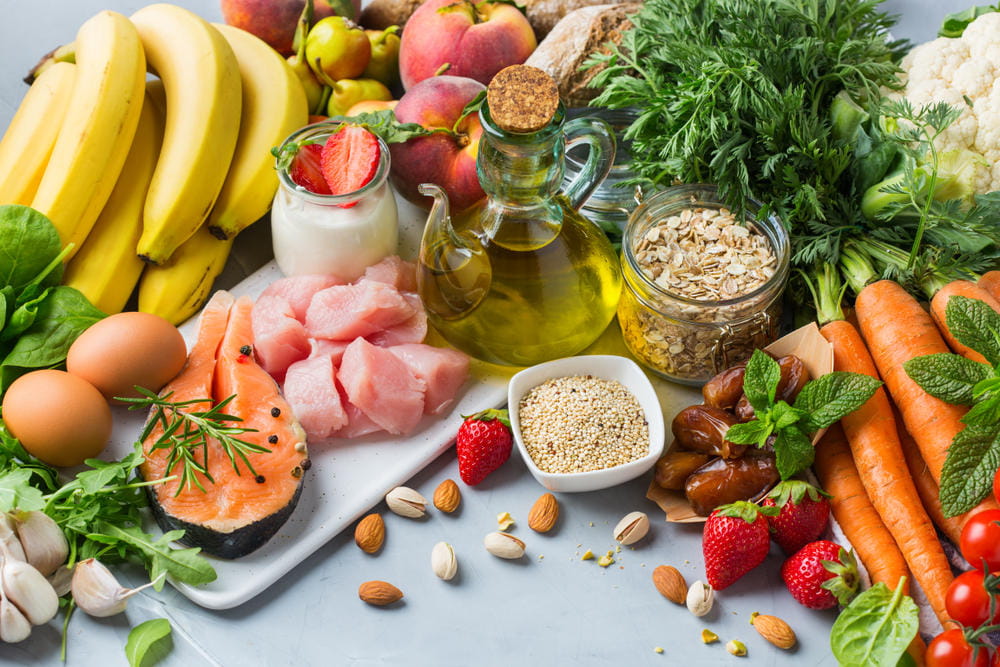 Healthy Mediterranean diet foods on a table, including bananas, eggs, salmon, lean chicken, yogurt, olive oil, seeds, nuts, and carrots