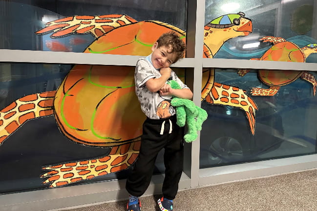 Smiling child hugging a stuffed turtle in front of a mural of a turtle