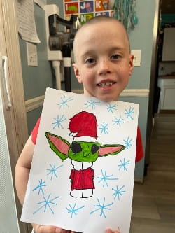 Smiling young boy holding drawing of Baby Yoda (Grogu) dressed as Santa. 