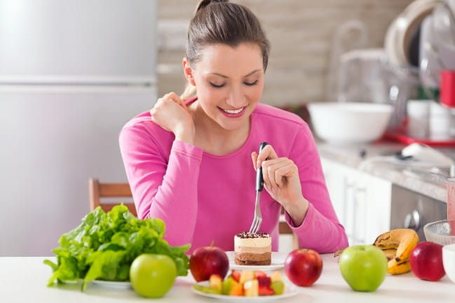 Person eating a processed dessert while surrounded by whole fruits and vegetables.