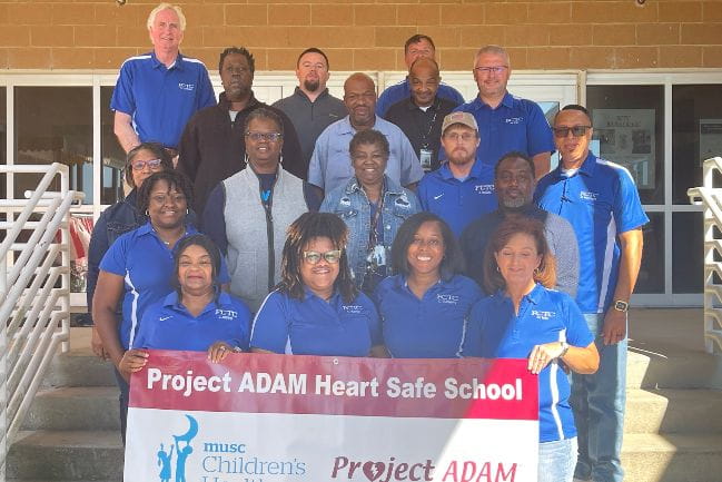 Group of smiling people holding a sign saying Project ADAm Heart Safe School