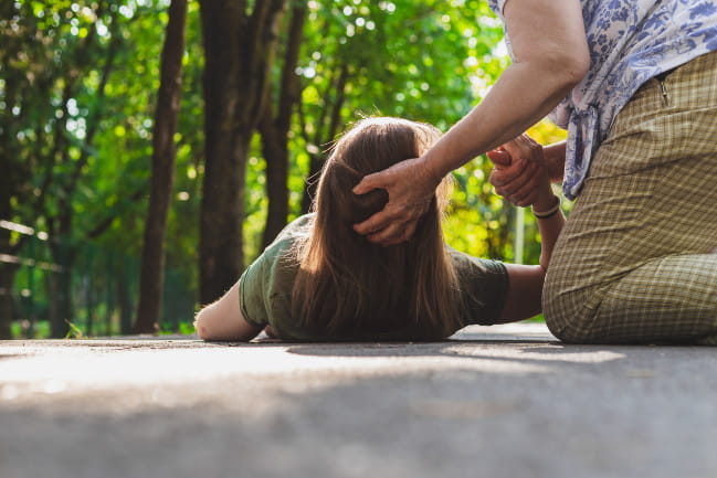 A person that has fallen on the ground outside, with another person knelt beside them and holding the fallen person's head up.