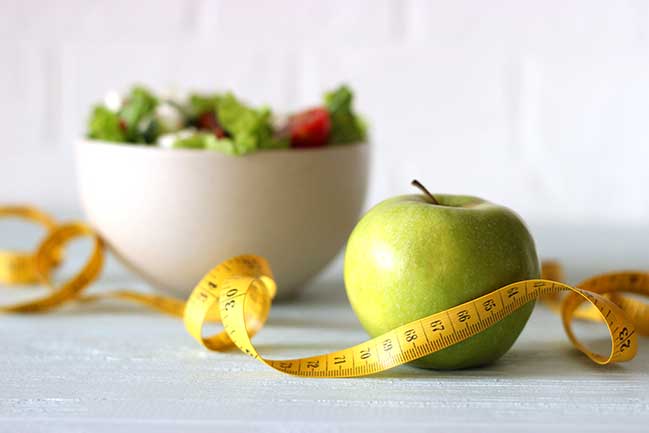 A green apple with a measuring tape wrapped around it as a bowl of salad sits in the background.