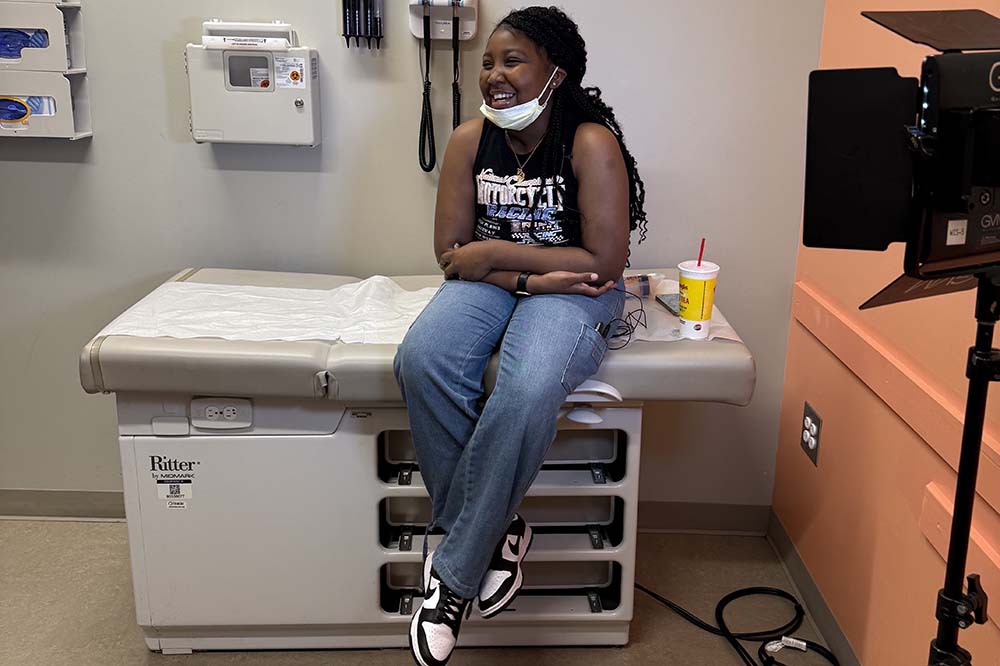 A girl sits on a clinic table. She is smiling and wearing a surgical mask, T-short, jeans and sneakers.